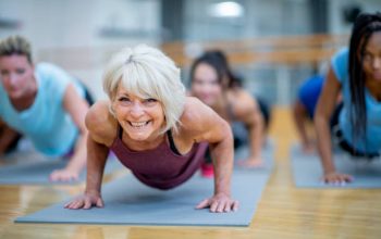 An older Caucasian woman is seen holding a plank pose while participating in a  co-ed, multi-ethnic, fitness class.  She is smiling and appearing to enjoy the class.